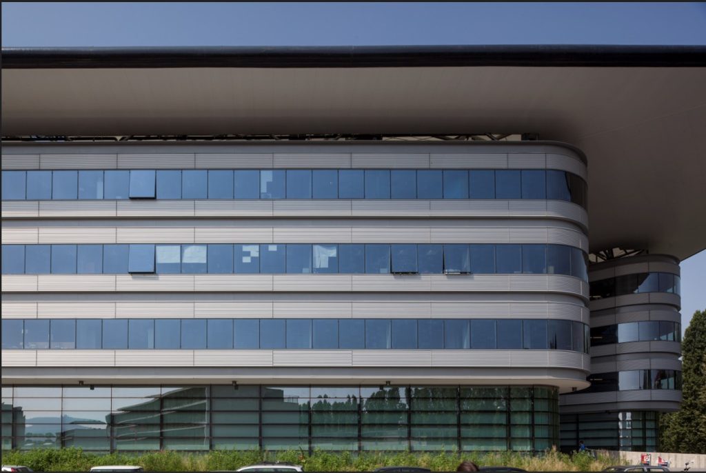 Facciata moderna del Campus Luigi Einaudi, con fasce orizzontali di vetrate blu, angoli arrotondati e un’ampia copertura sospesa.