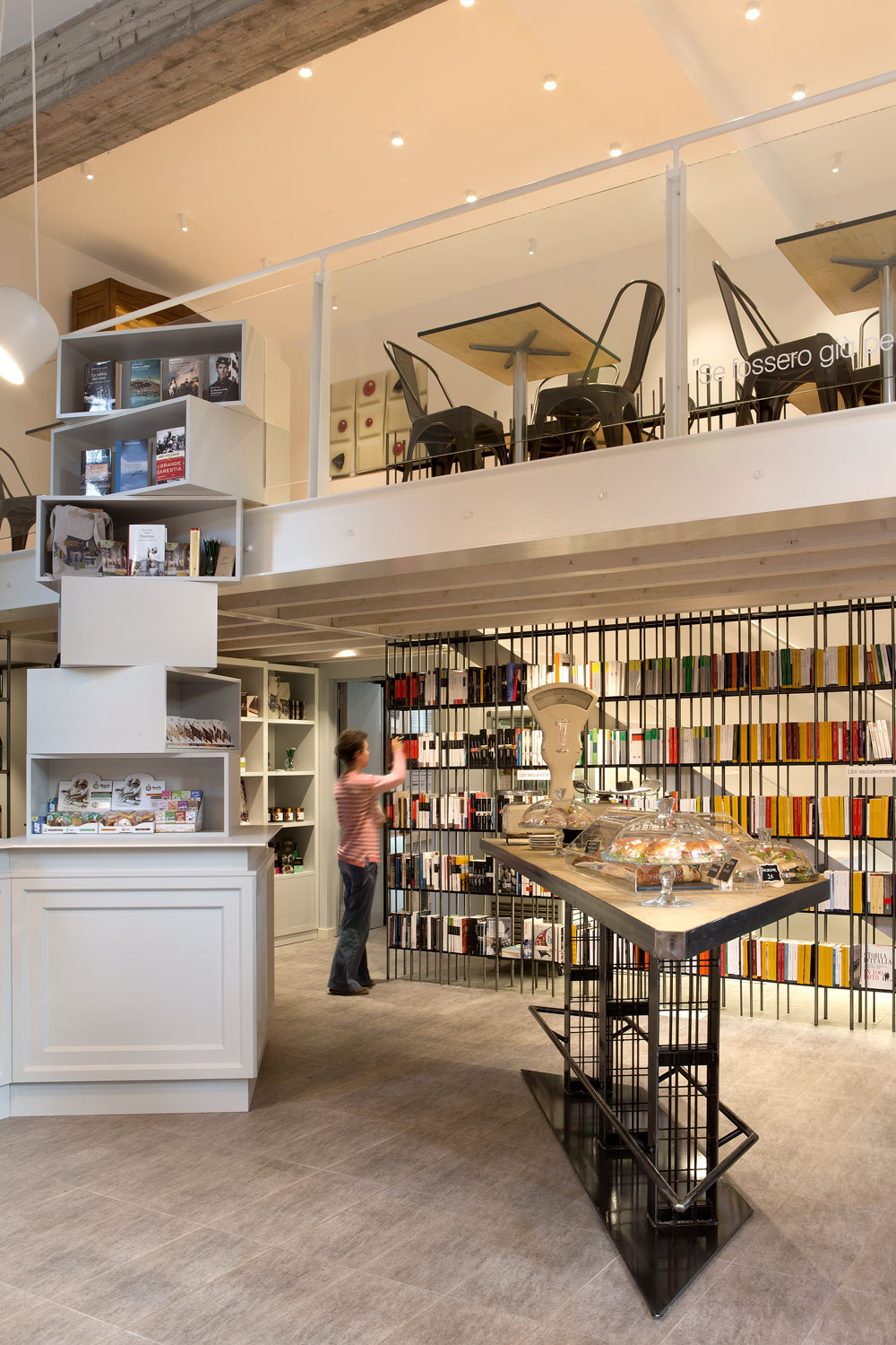 Interno di una libreria-caffetteria su due livelli, con scaffali pieni di libri, tavolo centrale con dolci esposti e una persona che sistema i volumi.
