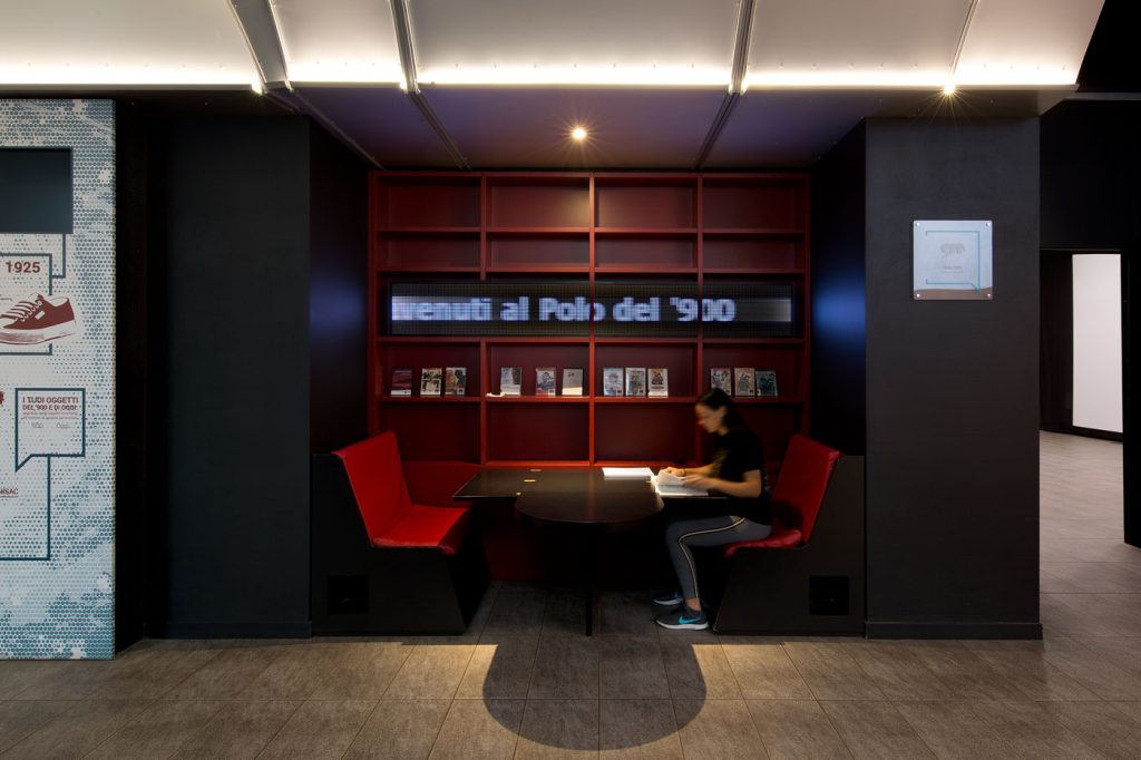 Area lettura del Polo del ’900 con sedute rosse, scaffale di libri e una persona seduta al tavolo; sullo sfondo, la scritta luminosa “Benvenuti al Polo del ’900”.