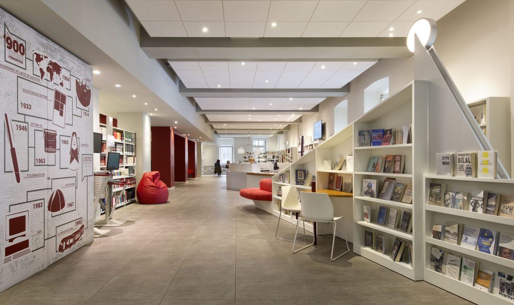 Corridoio di una biblioteca moderna con scaffali di libri, postazioni di lettura e sedute rosse; sulla parete sinistra, una cronologia illustrata del Novecento piemontese.