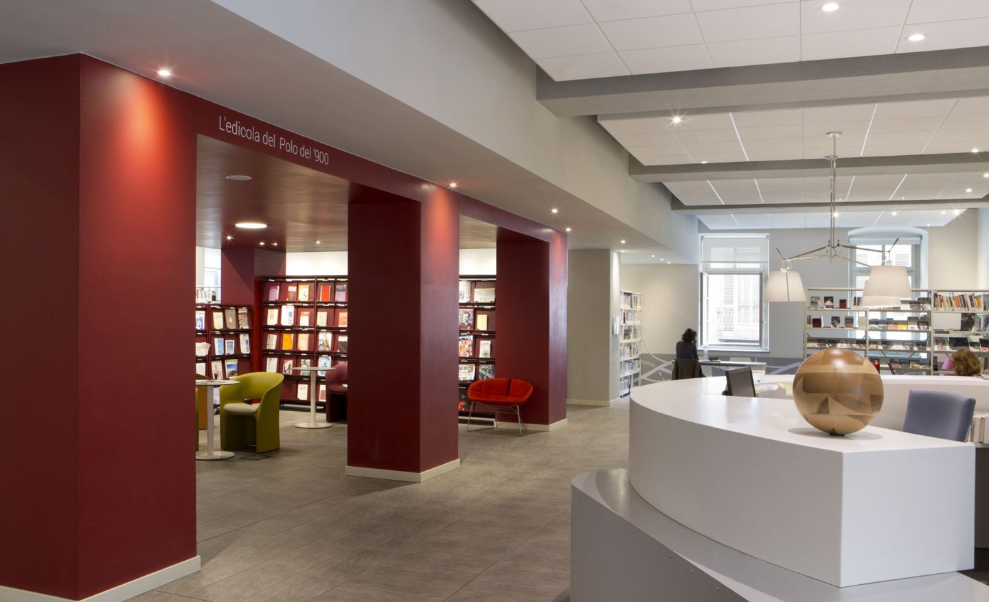 Interno moderno di una biblioteca, con banco informazioni circolare in primo piano e area lettura “L’edicola del Polo del ’900” incorniciata da pareti rosse, scaffali di libri e poltrone colorate.
