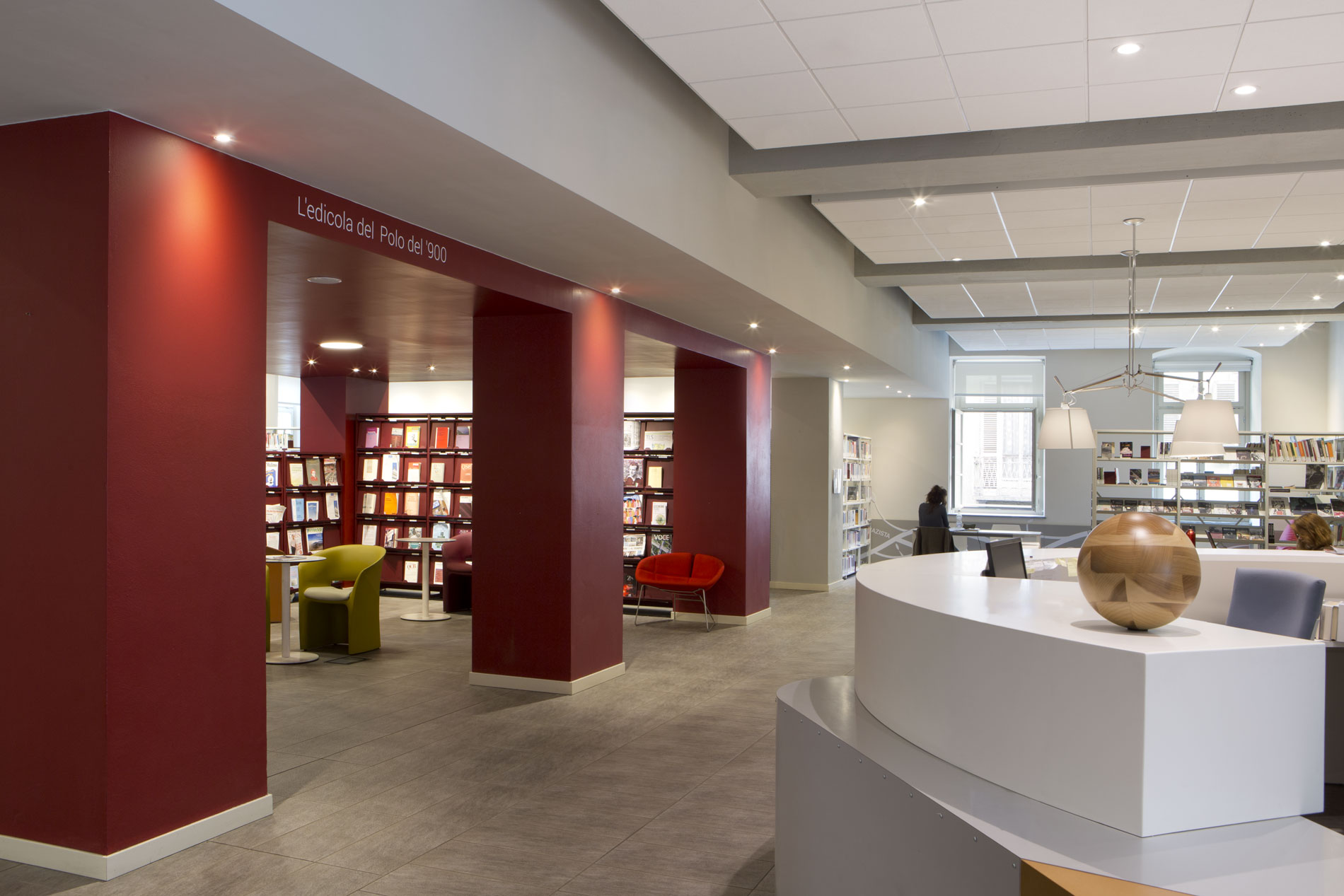 Interno moderno di una biblioteca, con banco informazioni circolare in primo piano e area lettura “L’edicola del Polo del ’900” incorniciata da pareti rosse, scaffali di libri e poltrone colorate.