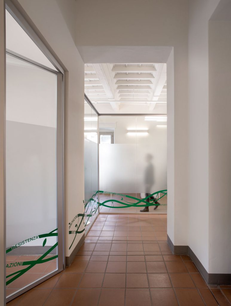 Corridoio luminoso con pareti vetrate satinate decorate da linee verdi e parole legate alla storia sociale; dietro il vetro si intravede la sagoma sfocata di una persona.
