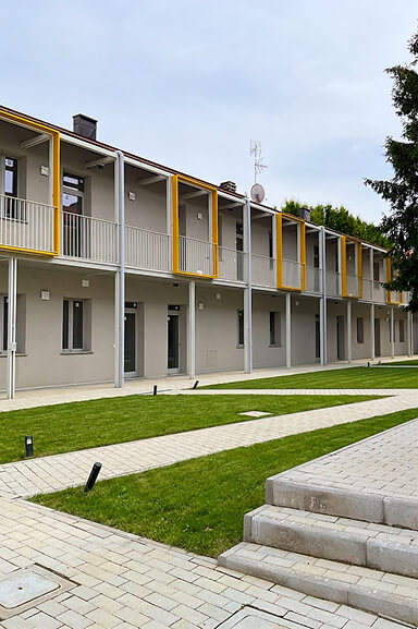 Vista laterale degli alloggi affacciati sulla corte, con ballatoi, cornici gialle, giardino e percorsi pavimentati.