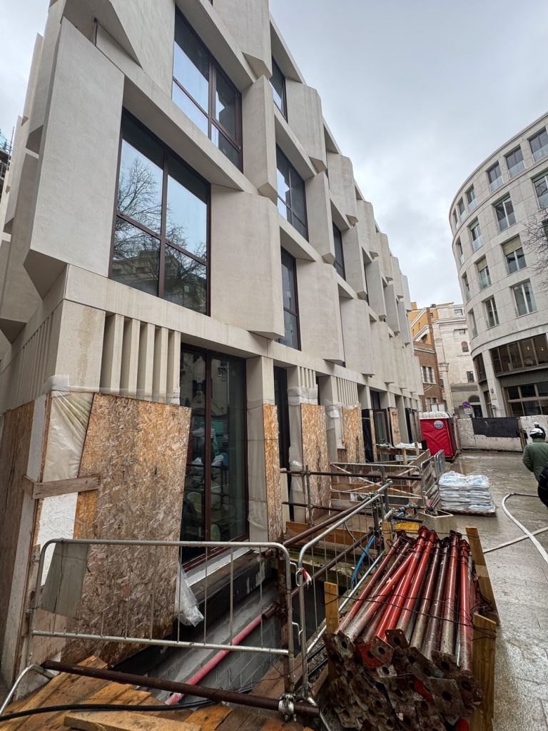 Vista dal basso della facciata geometrica in pietra chiara di un edificio moderno in cantiere, con puntelli metallici e pannelli protettivi in primo piano.