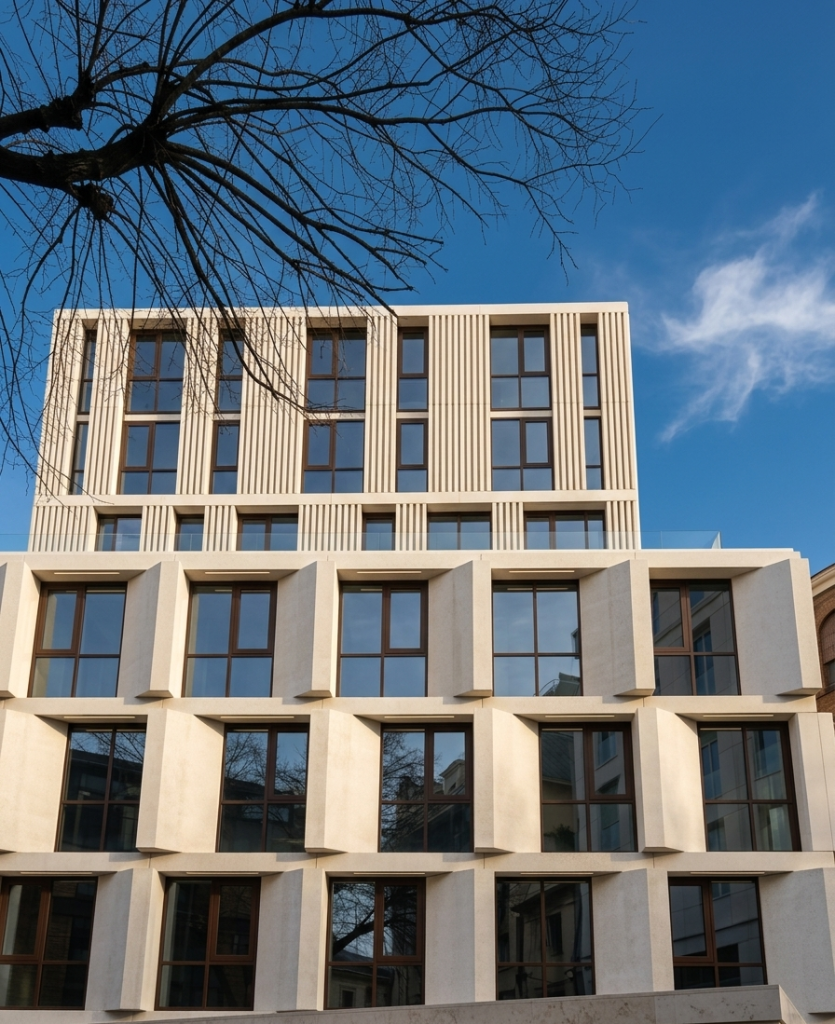 Vista dal basso della facciata in pietra chiara di un edificio moderno con moduli geometrici sporgenti attorno alle finestre e scanalature verticali nei piani superiori, sotto un cielo azzurro.