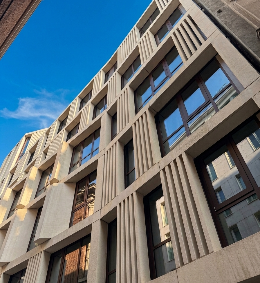 Prospettiva dal basso della facciata in pietra chiara di un edificio moderno, con scanalature verticali e ampie finestre, sotto un cielo azzurro.