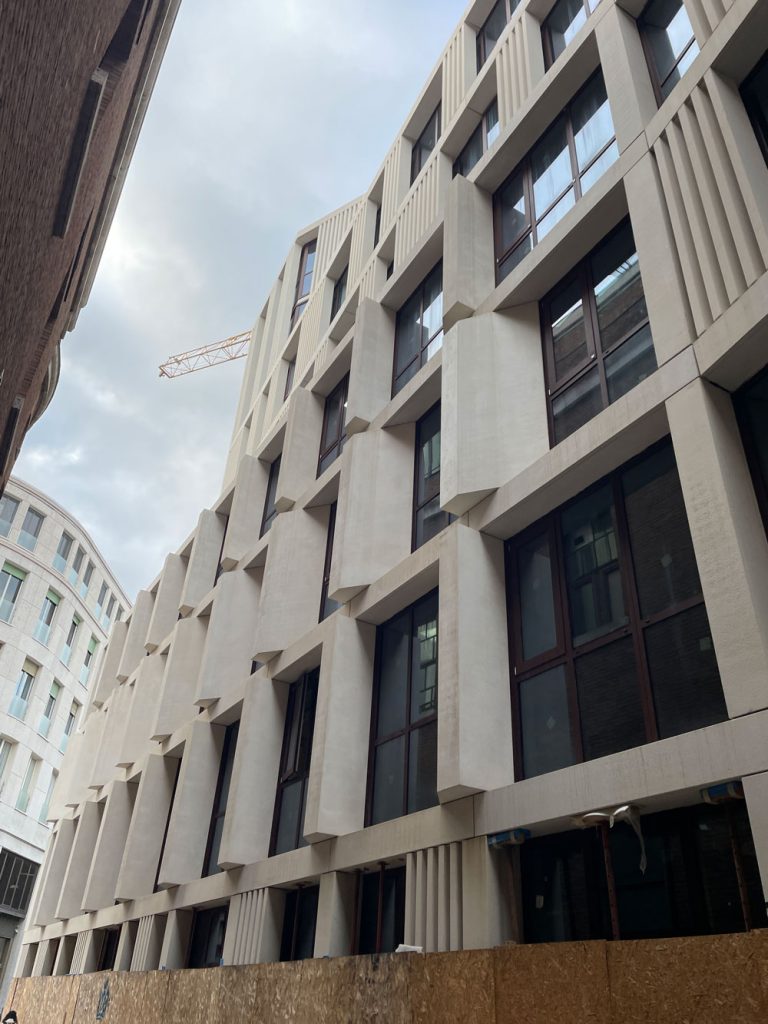 Vista dal basso della facciata geometrica in pietra chiara di un moderno edificio, con sporgenze angolari attorno alle finestre e una gru edile visibile tra le strutture urbane.