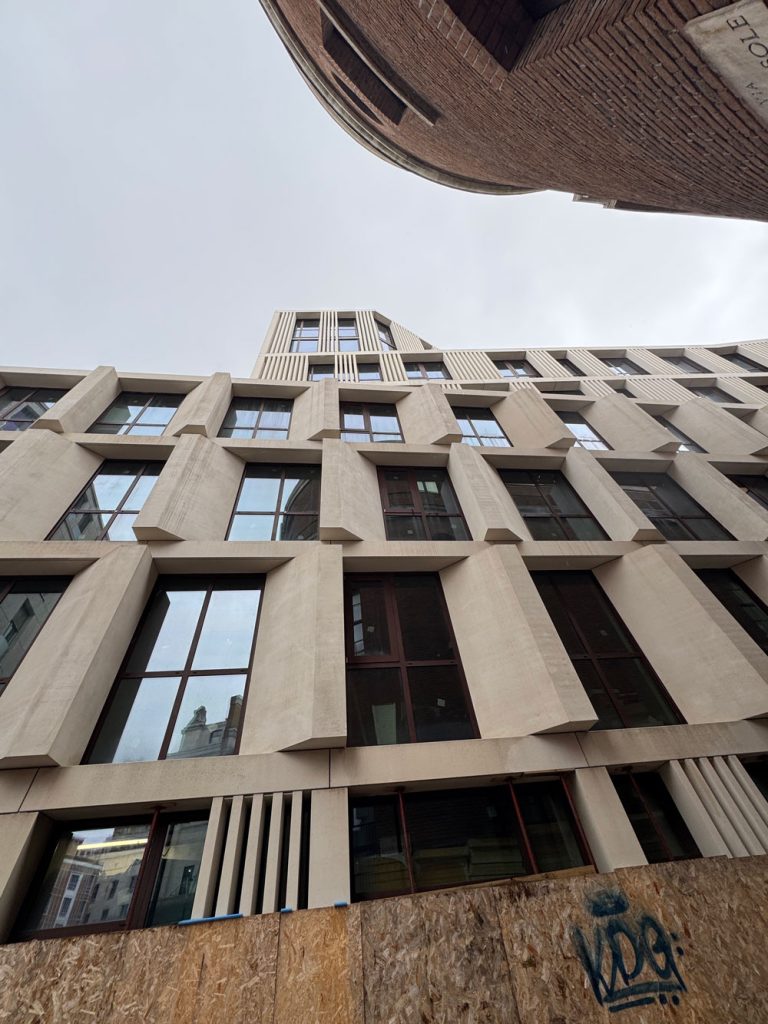 Inquadratura dal basso verso l'alto della facciata in pietra chiara di un moderno edificio, caratterizzata da rilievi geometrici angolari attorno alle finestre.