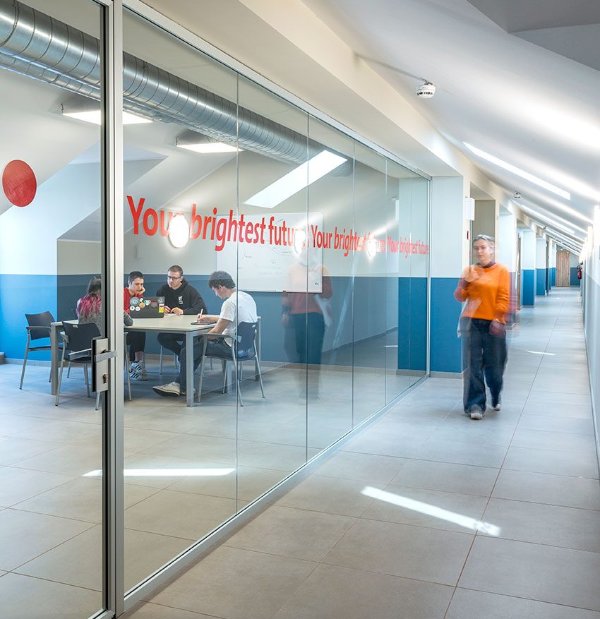 Corridoio moderno con parete di vetro che mostra quattro persone sedute a un tavolo in una sala riunioni e una quinta persona in movimento nel corridoio, con la scritta rossa "Your brightest future" sul vetro.