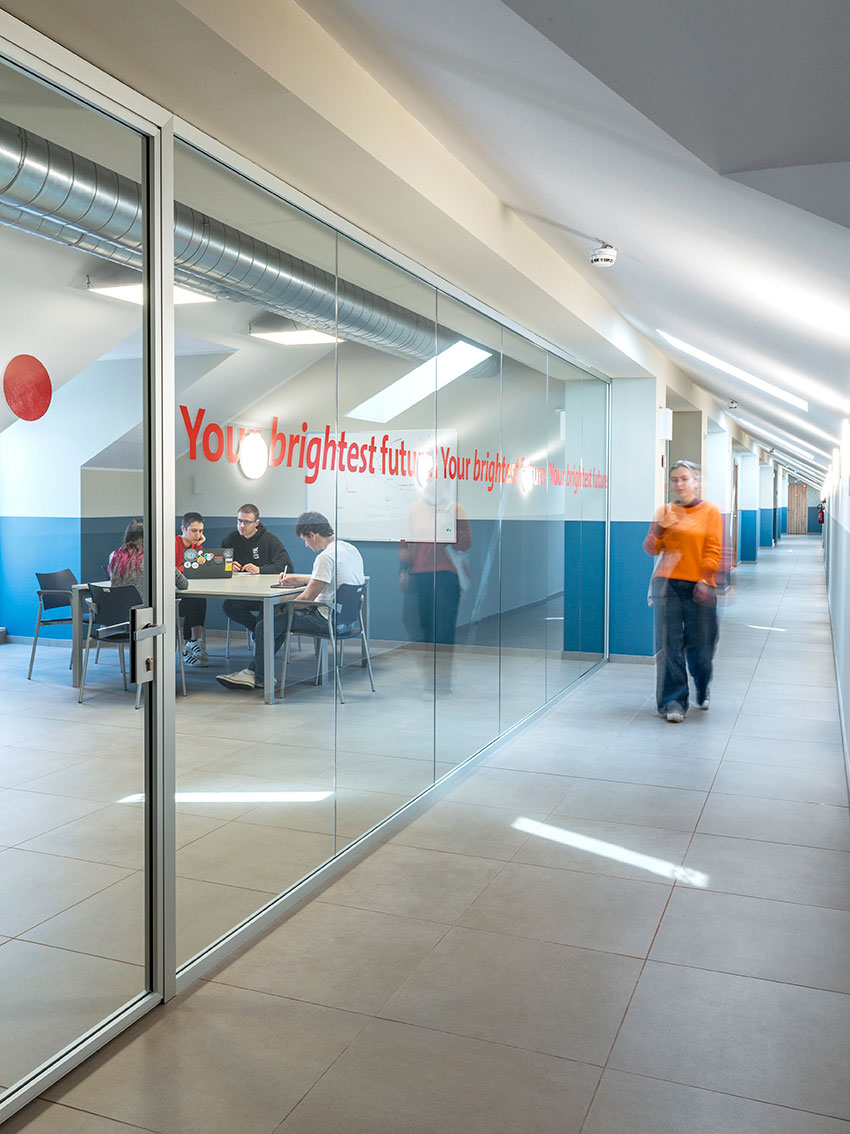 Corridoio moderno con parete di vetro che mostra quattro persone sedute a un tavolo in una sala riunioni e una quinta persona in movimento nel corridoio, con la scritta rossa "Your brightest future" sul vetro.