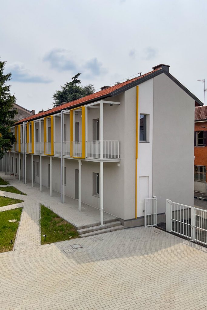 Vista laterale del complesso residenziale di Legnano, con ballatoio, cornici gialle, percorsi pavimentati e aree verdi interne.