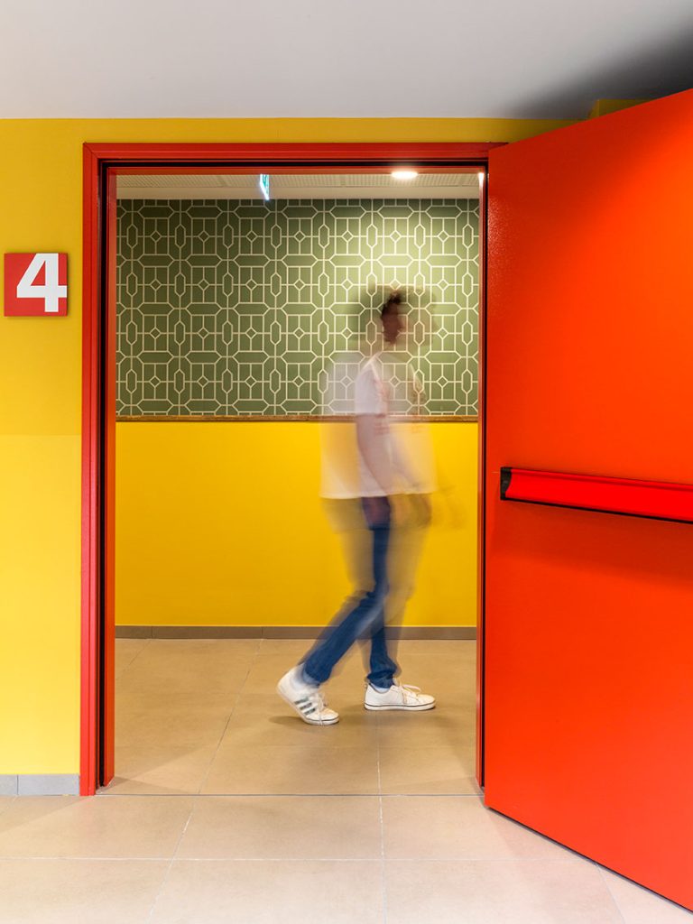 Una figura sfocata dal movimento indossa una t-shirt bianca e jeans mentre attraversa una porta rossa aperta, passando da un corridoio giallo con un cartello '4' rosso a una stanza con carta da parati verde a motivi geometrici e boiserie gialla.