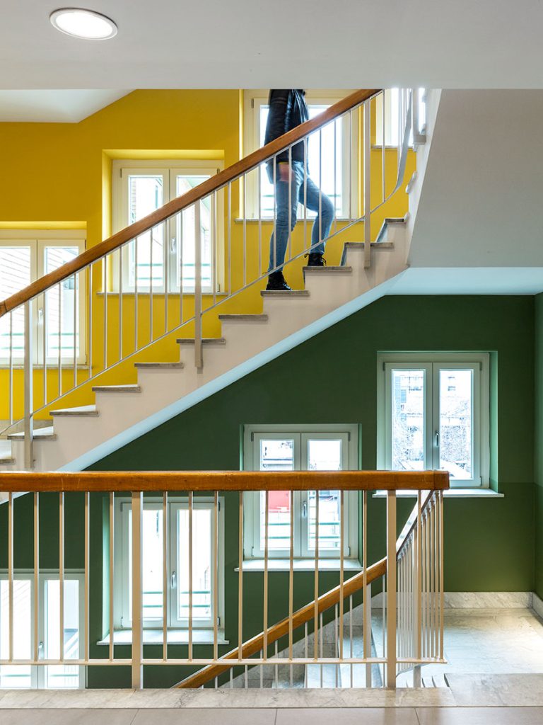 Una persona sale una scala a chiocciola in un moderno edificio con pareti colorate di giallo e verde e finestre.