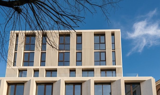 Facciata di un edificio moderno con finiture chiare, elementi verticali a listelli, ampie vetrate e rami spogli in primo piano contro un cielo azzurro.