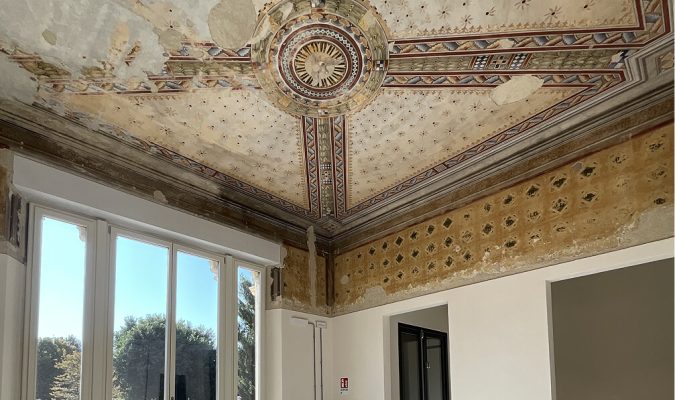 Soffitto d'epoca affrescato con rosette geometriche e motivi decorativi in una stanza in fase di ristrutturazione con ampie finestre luminose.