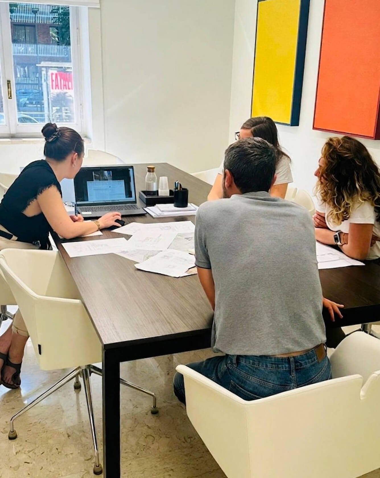 Gruppo di persone sedute attorno a un tavolo da riunione mentre consultano un laptop e documenti cartacei in un ufficio moderno.